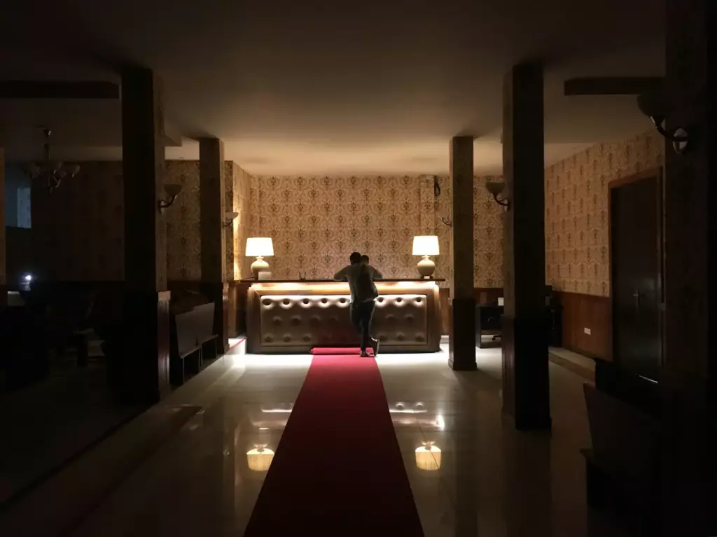 A man in a dimly lit hotel lobby asking the night porter for discreet advice about brothels in Lisbon.